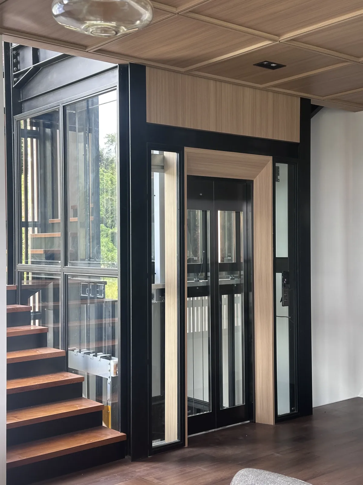 Glass home lift in a black-framed shaft with oak-veneer portal and soffit, beside an open timber stair in a contemporary house