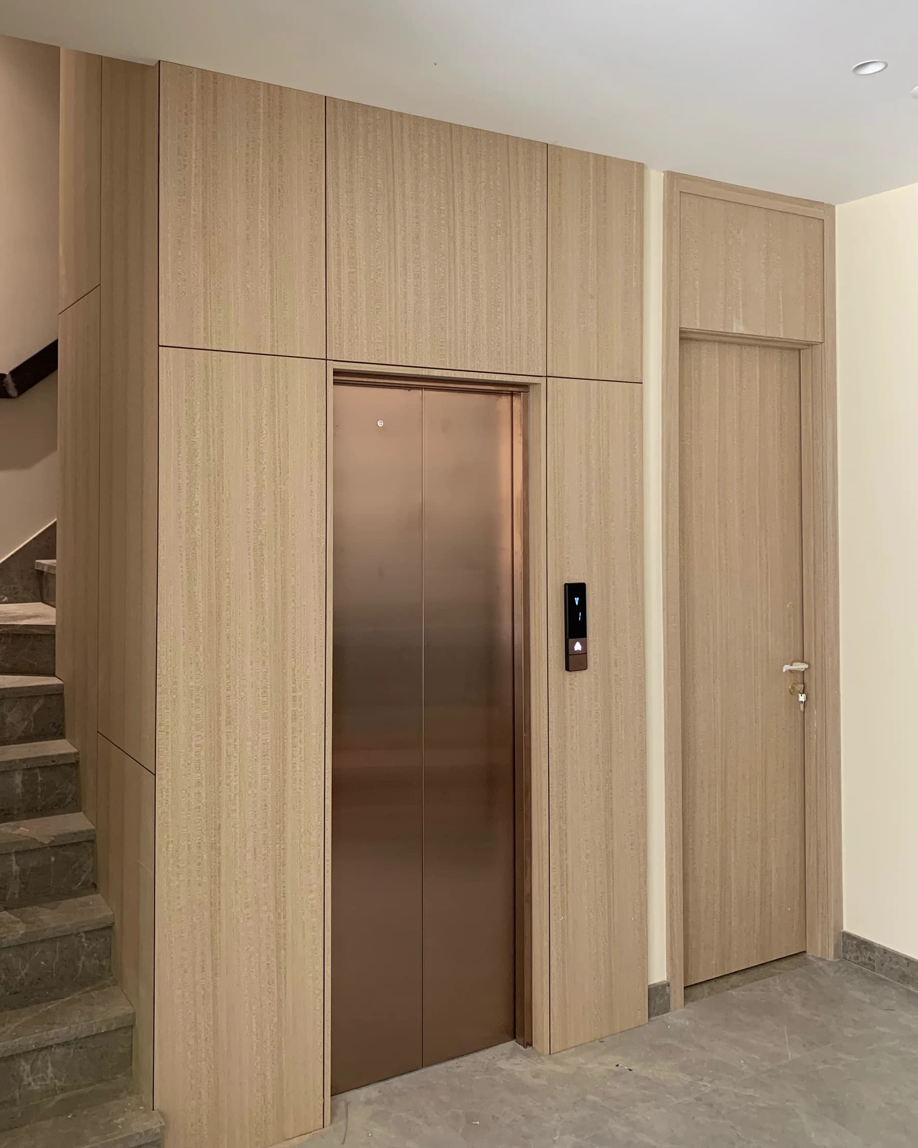 Tenkai V300 landing in a timber-panelled lobby, its bronze sliding doors closed, the black landing panel beside them and a stone stair alongside
